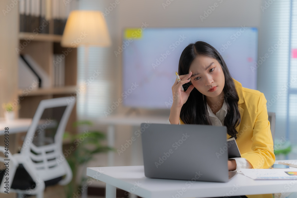 A young female employee of Asian descent, seated in her office, expresses weariness from the demands of her laptop-based work. A young female employee of Asian descent, seated in her office, expresses weariness from the demands of her laptop-based work.