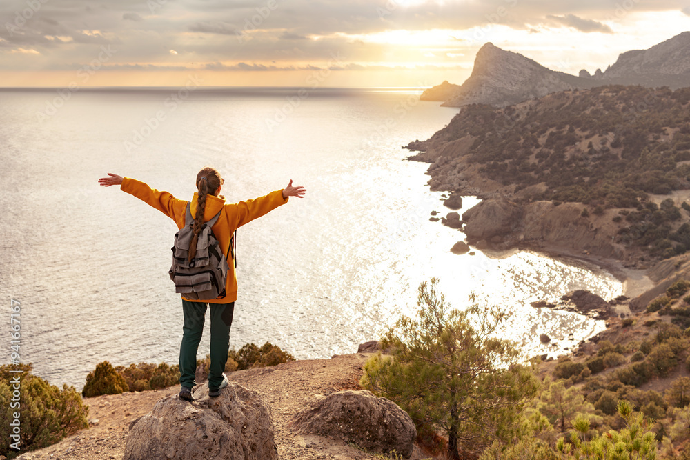 Happy young female hiker is standing with open arms at the cliff and enjoys sea view and sunset Happy young female hiker is standing with open arms at the cliff and enjoys sea view and sunset