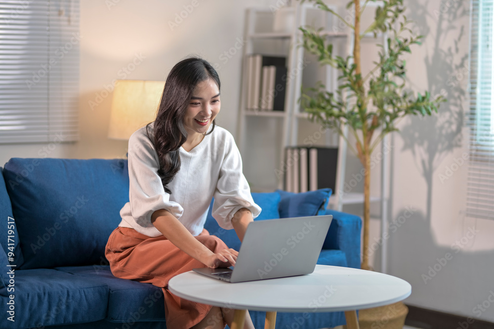 Young asian woman smiling and typing on laptop while working from home on sofa Young asian woman smiling and typing on laptop while working from home on sofa