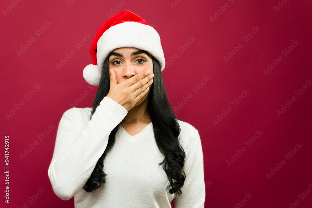 Shocked young woman in a santa hat stands in front of a red background, covering her mouth with her hand Shocked young woman in a santa hat stands in front of a red background, covering her mouth with her hand