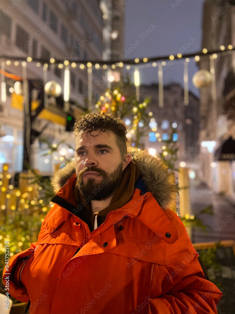 Fototapeta premium Handsome millennial man model with a hairstyle in winter red jacket walks in night city with festive lights. Christmas and winter holidays season