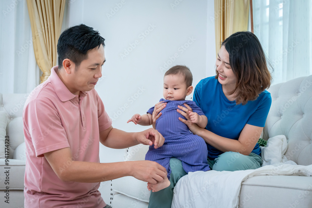 A man and a woman are sitting on a couch with a baby in between them. The baby is wearing a purple outfit. Scene is warm and loving, as the family is spending quality time together