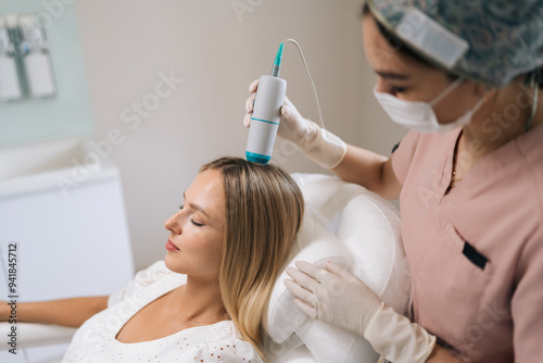 Closeup of trichologist in gloves and mask performing hair treatment procedure using spectrum phototherapy device. Blonde female patient having hair light therapy with modern innovation equipment.