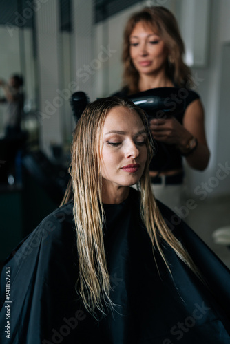 Vertical shot of female hairstylist drying and straightening long blonde customer hair with comb and hairdryer in beauty salon. Pretty young woman lady client getting stylish hairdo done at salon.