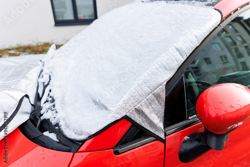 Close-up detail red car with snow-covered windshield protected by reflective cover mat, parked outside residential building during winter. Windscreen window ice frost protection photo