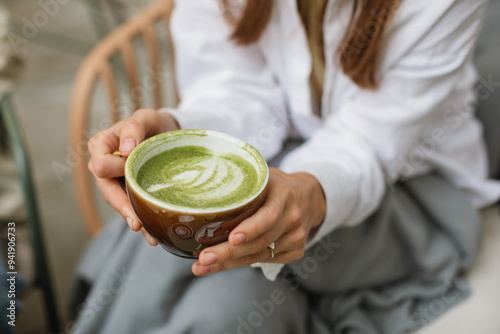 A close-up of a young woman holding a cup of matcha latte sitting on a cafe terrace.