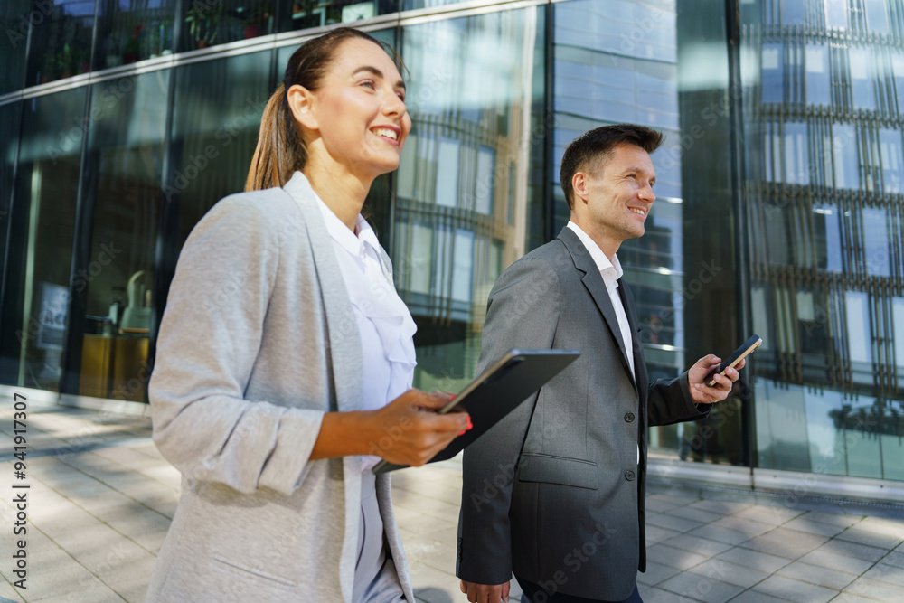 Two business professionals share a lighthearted moment while discussing work on a digital tablet outdoors. Two business professionals share a lighthearted moment while discussing work on a digital tablet outdoors.