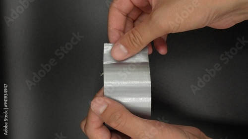 hands carefully peeling a strip of silver duct tape from a roll. The tape is being pulled away smoothly, highlighting the texture and sheen of the adhesive material. The minimalist setting, with its