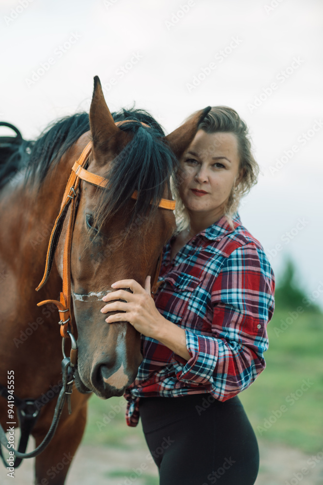 Obraz premium Happy blonde with horse in forest. Woman and a horse walking through the field during the day. Dressed in a plaid shirt and black leggings.