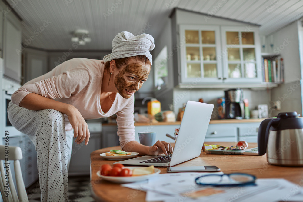 Woman with face mask enjoying breakfast and coffee while working on laptop in kitchen