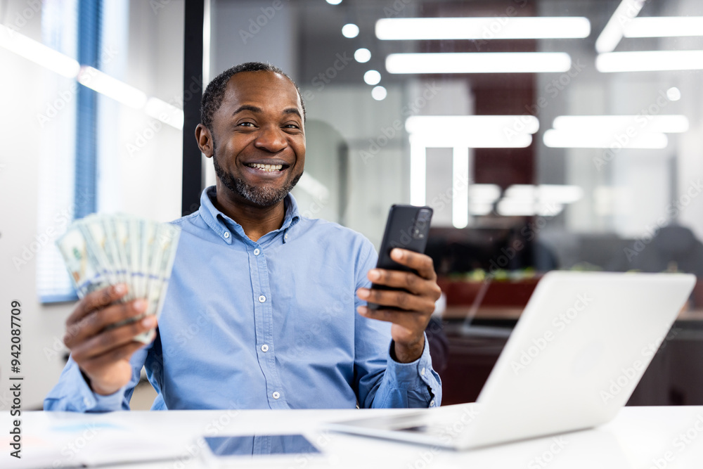 Confident man sitting in modern office, smiling while holding cash and smartphone. Demonstrates financial success, positive emotions, and work-life balance.