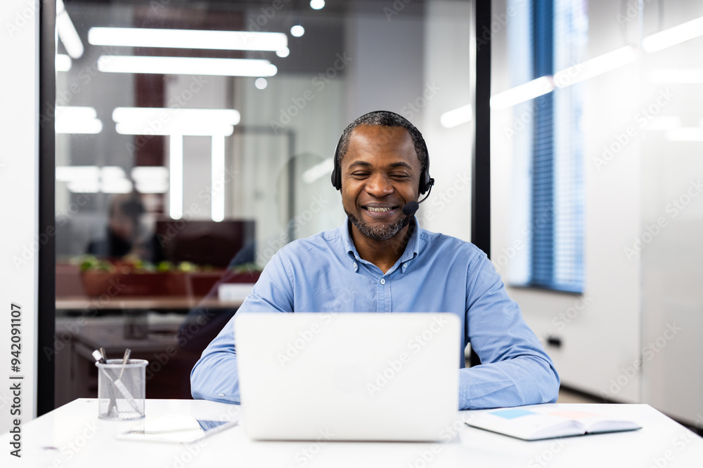 Confident businessman wearing headset engaged in online conversation on laptop. Smiling and communicating during virtual meeting in modern office setting. Concepts of technology, work, communication