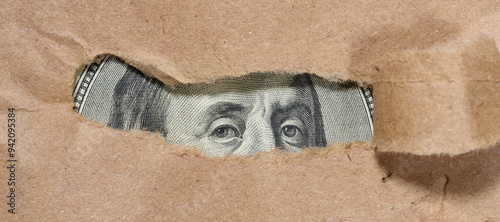 A piece of a hundred dollar bill is visible through a hole in the paper. Symbolism of hidden or concealed wealth, money that remains out of sight, or financial resources.