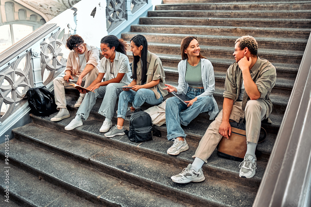 Fototapeta premium A group of student friends are sitting on the steps of the university inside and talking, getting ready for classes. Modern students.