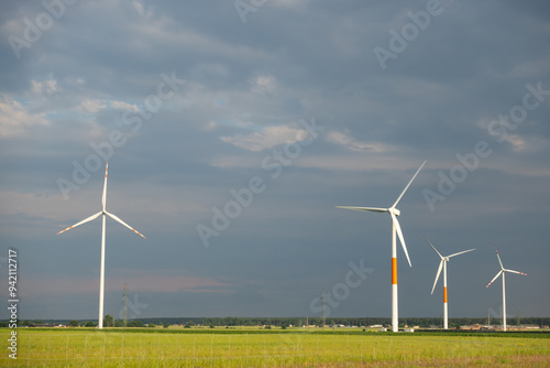 Obraz na płótnie Distant white wind turbines against a dramatic cloudy sky. Meadow in the foreground, taken late afternoon w salonie