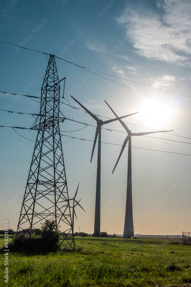 Wind turbines and high voltage electric pylon against blue sky. Sun shining behind the turbines
