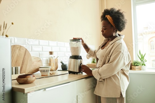 Side view of curly Black woman using food blender mixing ingredients for milk smoothie while cooking healthy breakfast at beige sunlit kitchen, copy space