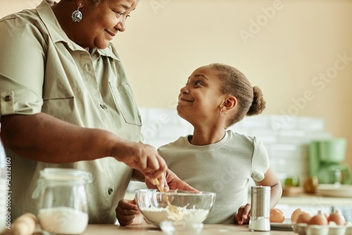 Side view of charming Black girl looking at loving grandma with adoring smile during cooking pancakes together at beige kitchen, copy space