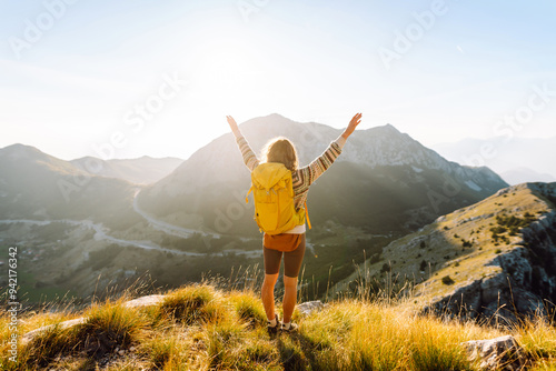 Cheerful young woman sits on a grassy slope, smiling as the sun sets behind the mountains. She has a backpack beside her and is taking a moment to enjoy the beautiful landscape. Lifestyle, tourism.