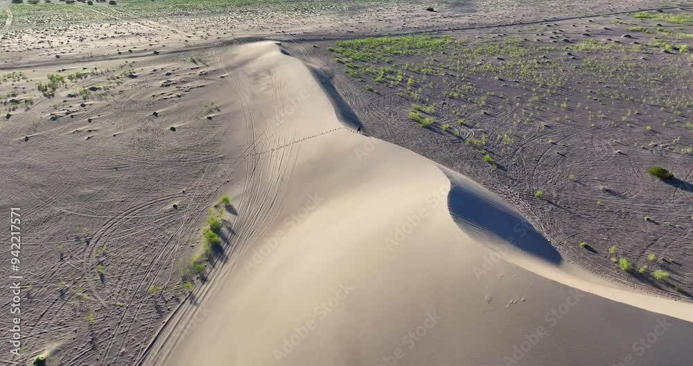 Drone shot of a man walking up a desert sand dune on its crest.