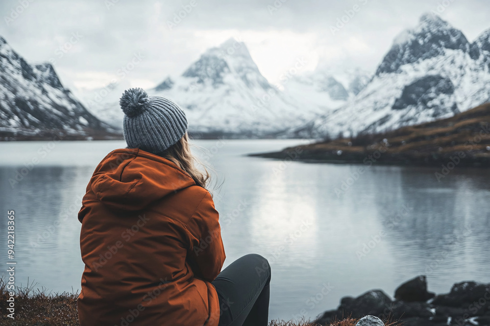 woman in an orange jacket and beanie sits by a mountain lake with snow-capped peaks creating a serene and reflective scene woman in an orange jacket and beanie sits by a mountain lake with snow-capped peaks creating a serene and reflective scene