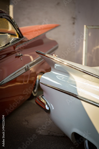 Zwei große amerikanische Coupe Autos als Lowrider Custom mit Speichenfelgen und Weißwandreifen in einem Industriegebiet. Detailaufnahme der Heckflossen bei Sonnenschein.