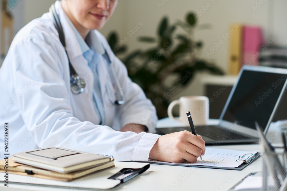 Medical professional wearing lab coat working at desk. Completing paperwork with pen, stethoscope around neck, book, laptop, and coffee mug on desk Medical professional wearing lab coat working at desk. Completing paperwork with pen, stethoscope around neck, book, laptop, and coffee mug on desk