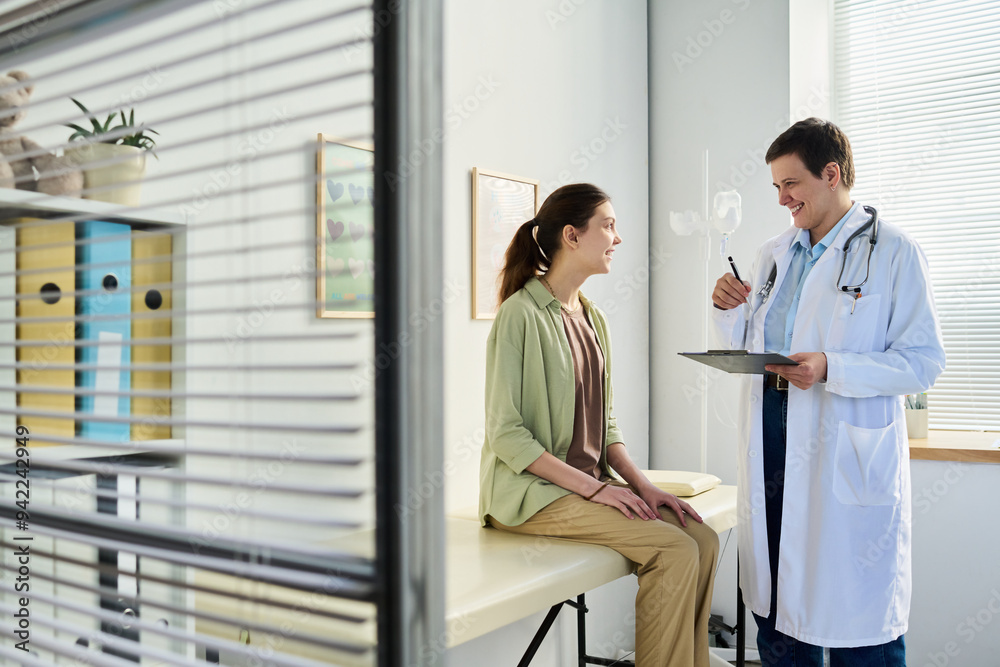 Obraz premium Medical professional in white coat discussing health with young patient sitting on examination table in modern consultation room with equipment and documents present