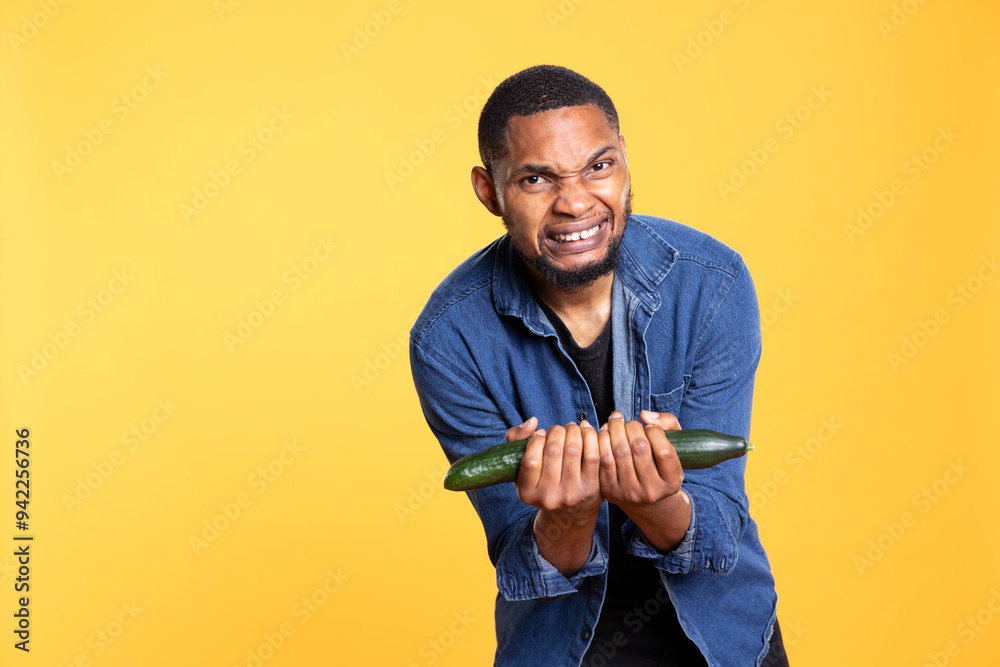 African american young guy fooling around with a cucumber, acting like he does weightlifting against yellow background. Motivated happy adult having fun with fresh bio veggies. African american young guy fooling around with a cucumber, acting like he does weightlifting against yellow background. Motivated happy adult having fun with fresh bio veggies.