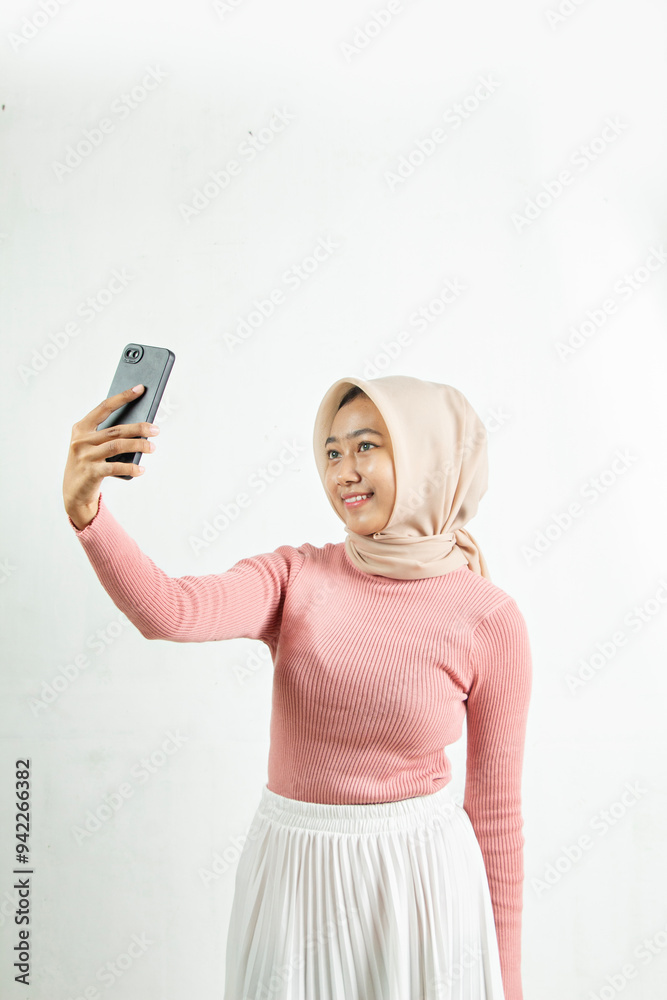 Naklejka premium Advertisement The Muslim teenager took a selfie with a cellphone with a white background. The woman was wearing a hijab and a pink woven dress and white skirt.