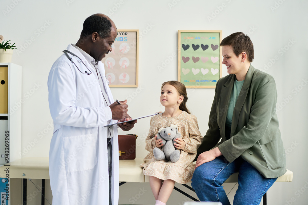 © pressmaster - Child sitting on exam table holding teddy bear while doctor talks to parent