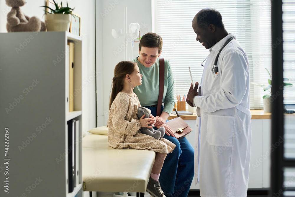 Male doctor standing near seated child and mother in clinic room. Child holding stuffed toy while doctor explaining something to them Male doctor standing near seated child and mother in clinic room. Child holding stuffed toy while doctor explaining something to them
