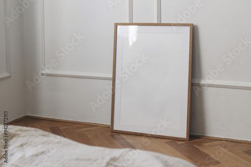 Vertical thin picture frame mount mockup. Empty wooden frame poster mock-up template on oak wooden parquet floor. White wall background, stucco decor. Minimal Scandinavian interior, blurred bed sofa