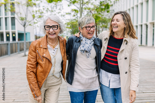 Three mature retired women laughing while walking together outside. Small group of 60s females having fun enjoying a conversation bonding at city street. Elderly friendship concept. photo