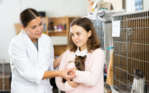 Cheerful cute tween girl standing with young female shelter worker near cages for homeless animals, hugging frightened little kitten photo