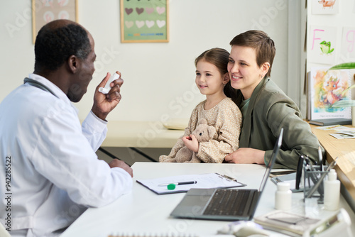 Doctor explaining medication to smiling mother and daughter in office, with medical notes and diagrams on the walls, nurturing a positive and friendly atmosphere photo