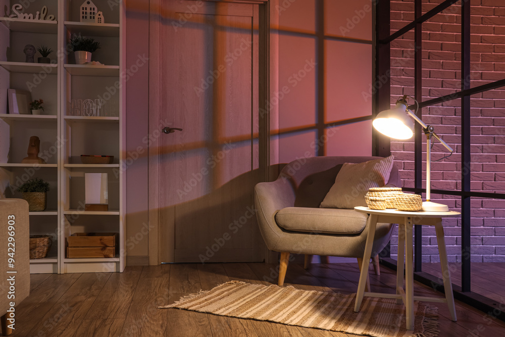 Fototapeta premium Comfortable armchair, table and glowing lamp in interior of living room