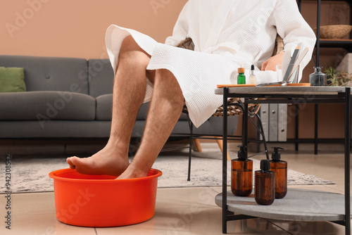 Handsome young man with tools keeping his legs in foot basin and doing pedicure at home, closeup photo