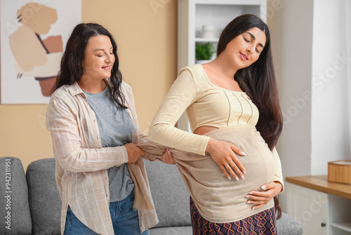 Happy doula massaging beautiful young pregnant woman with scarf at home