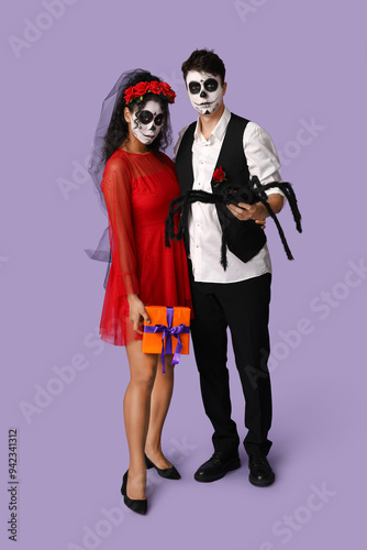 Young couple dressed for Halloween with toy spider and gift box on lilac background