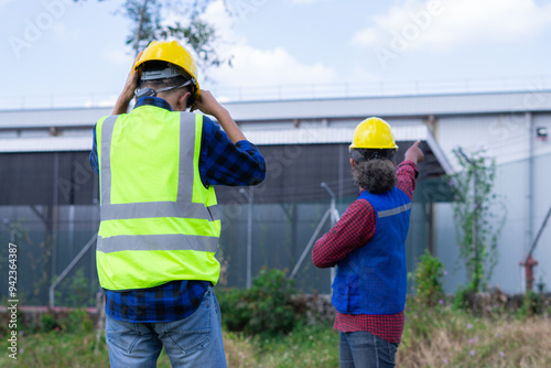 Two construction engineers in hard hats and safety vests collaborating on an industrial project, pointing at a building during a site inspection and planning.
