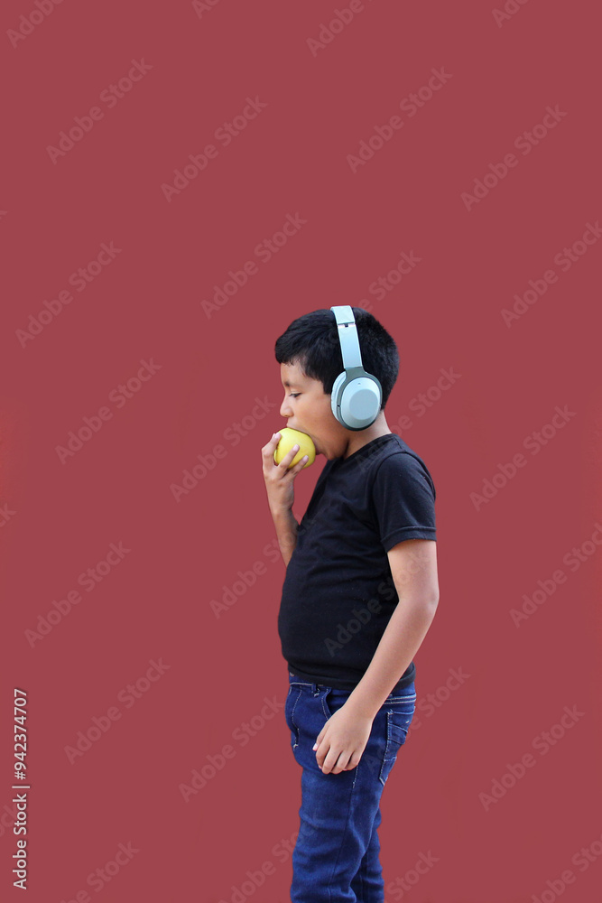 Obraz premium 9-year-old dark-skinned Latino boy with headphones eats an apple as a sign of healthy eating while listening to music