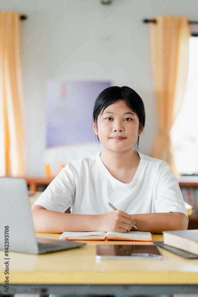 A young student sitting at a desk with a laptop, books, and a notebook, studying in a bright room with yellow curtains. A young student sitting at a desk with a laptop, books, and a notebook, studying in a bright room with yellow curtains.
