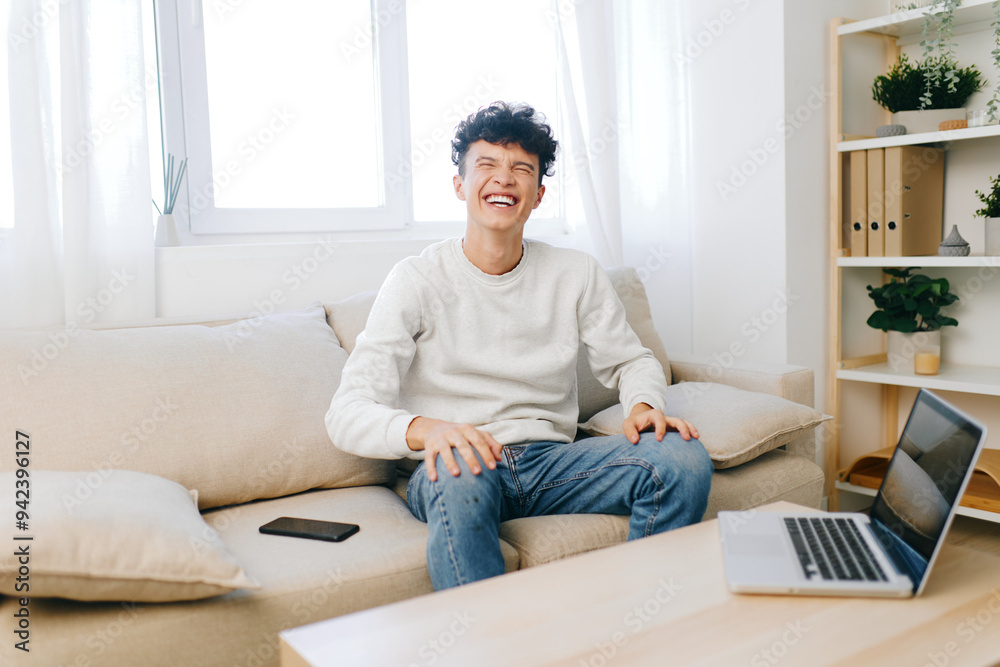 Young man smiling while sitting on a couch in a bright, airy room with a laptop nearby Natural light enhances the cozy atmosphere with indoor plants Young man smiling while sitting on a couch in a bright, airy room with a laptop nearby Natural light enhances the cozy atmosphere with indoor plants