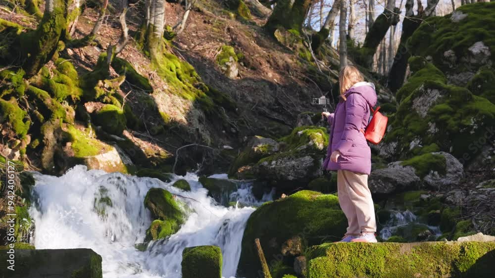 A woman stands peacefully by a flowing, picturesque waterfall, surrounded by vibrant green moss and tall trees