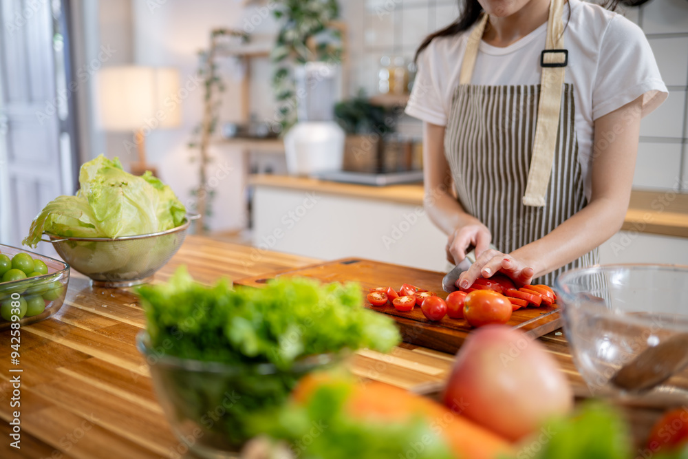 A cropped image of a woman slicing a tomato on a wooden cutting board at the kitchen table. A cropped image of a woman slicing a tomato on a wooden cutting board at the kitchen table.