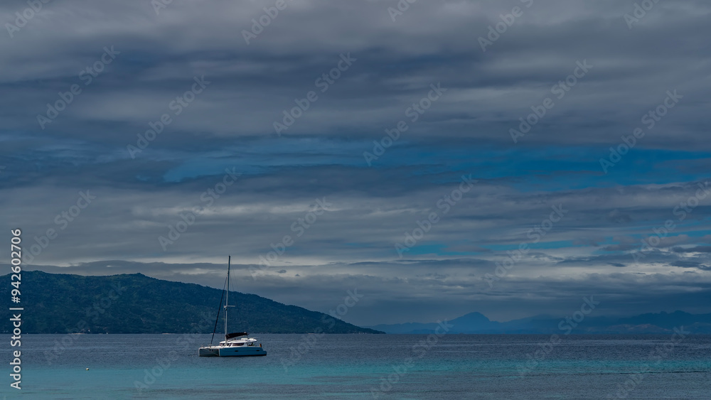 A lone white yacht is anchored in the turquoise ocean. A tall mast against a background of blue sky, clouds and mountains. A lone white yacht is anchored in the turquoise ocean. A tall mast against a background of blue sky, clouds and mountains.