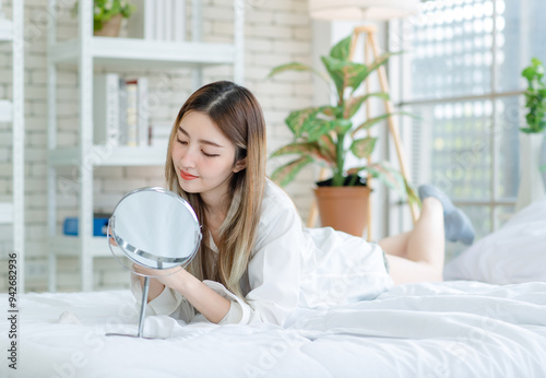 Asian young woman beautiful female happy smiling enjoying looking at the mirror with decorative makeup, beauty products, and cosmetics while lying on a white bed, Concept of teenage lifestyle at home. photo