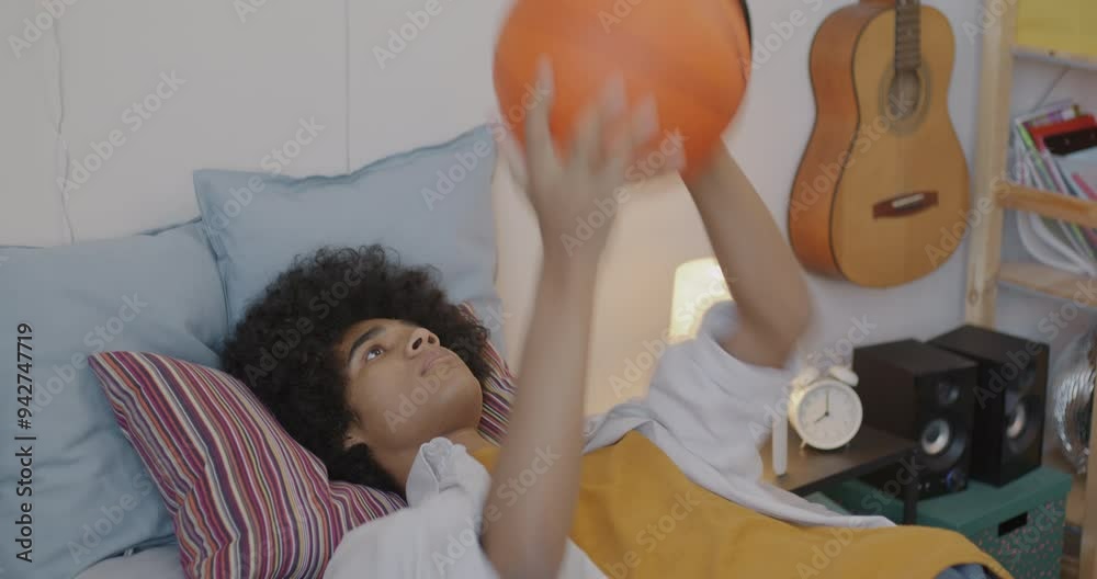 Bored African American teenager lying in bed throwing and catching basketball spending time at home. Leisure activity and teenage lifestyle concept.