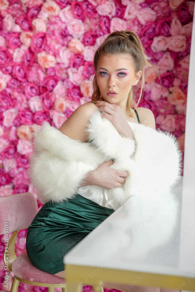 A beautiful girl in a green dress and a white fur coat poses in a studio. She stands in front of a wall of pink flowers. A beautiful girl in a green dress and a white fur coat poses in a studio. She stands in front of a wall of pink flowers.